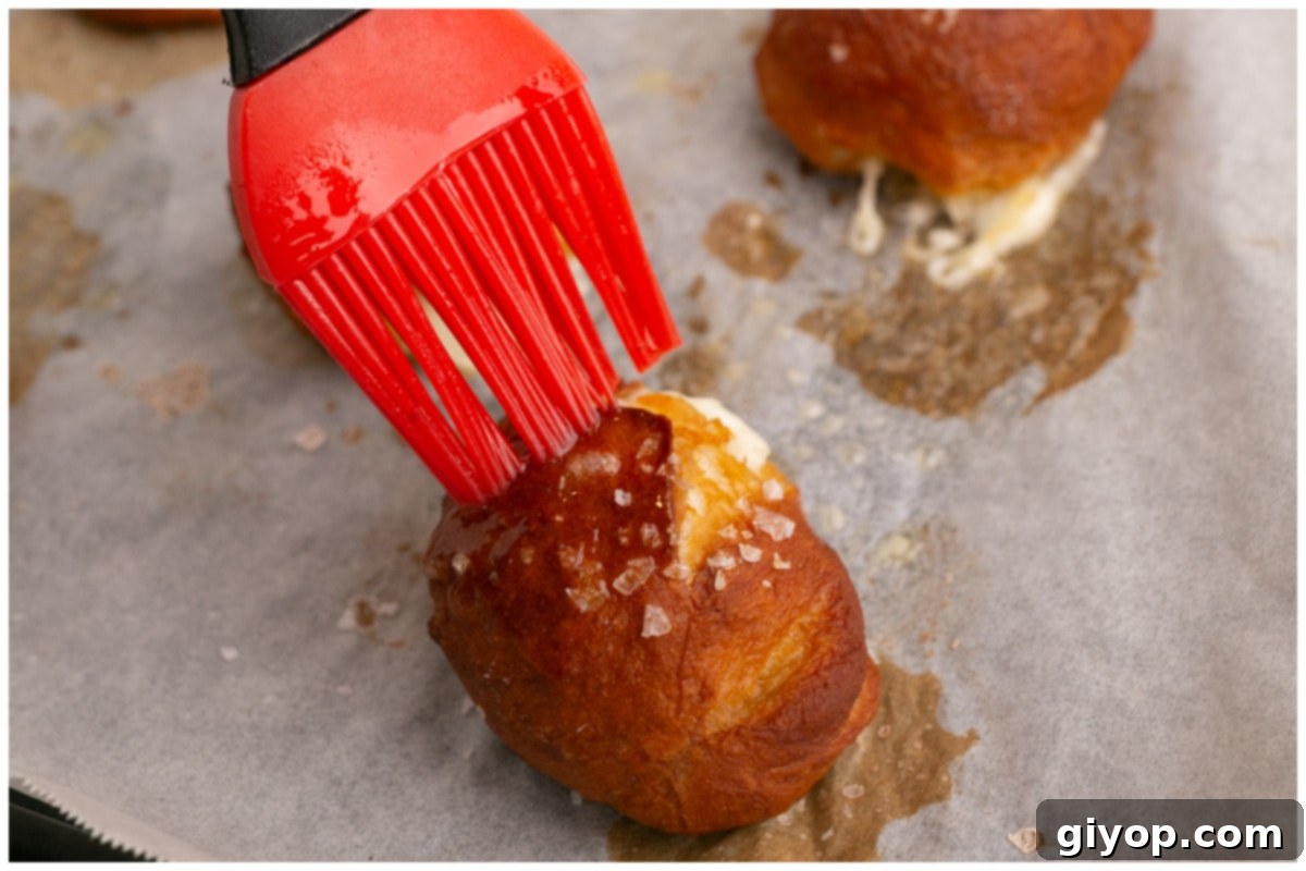 A hand brushing melted butter over hot, golden-brown pretzel bites on a baking sheet, adding a rich finish.