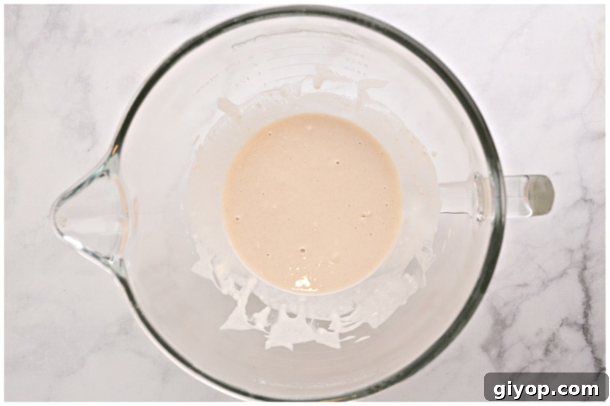 A glass bowl of a stand mixer containing the initial yeast, water, sugar, and flour mixture, beginning to proof.