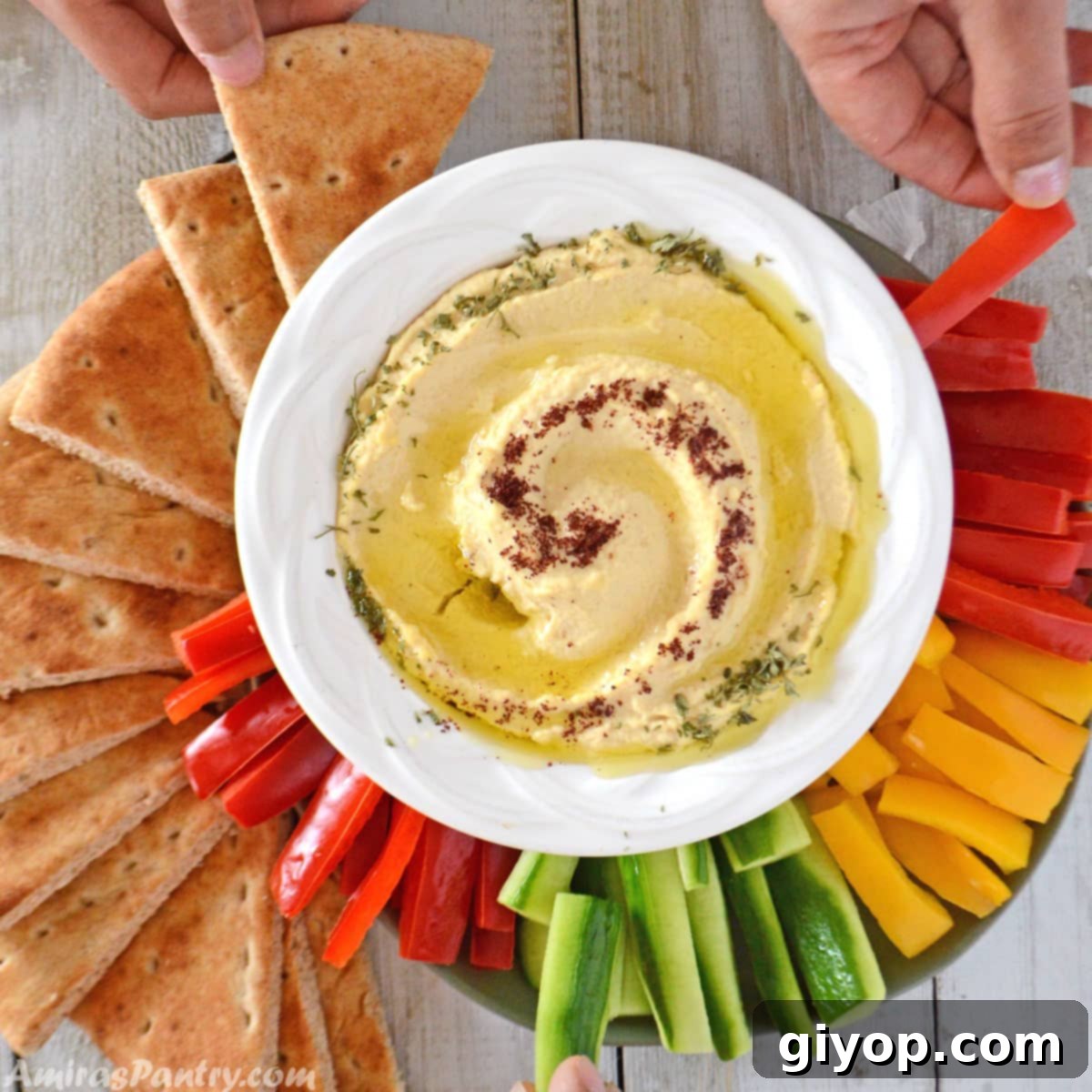 Hands reaching out to fresh vegetables with a large bowl of creamy hummus at the center of a rustic wooden table, inviting healthy snacking.