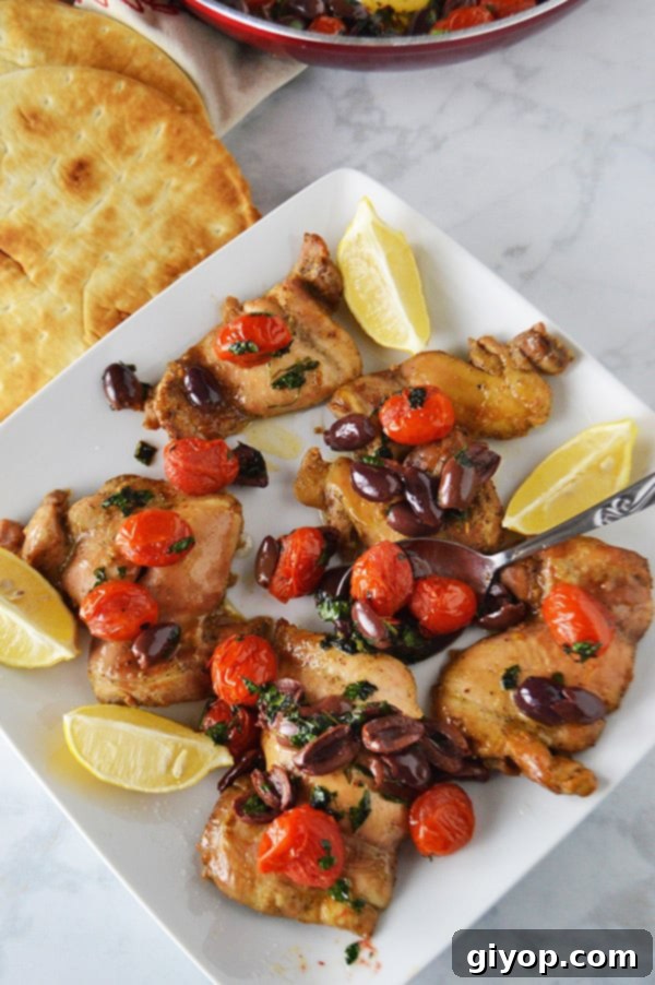 A large baking dish filled with beautifully baked lemon pepper chicken pieces, glistening with olive oil and adorned with a colorful topping of black olives and roasted tomatoes. Lemon wedges are scattered on the side, next to warm pita bread.