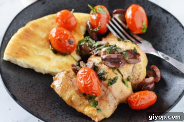 A beautifully plated serving of baked lemon pepper chicken in a black dish, adorned with the rich, dark Kalamata olive and roasted tomato relish. A piece of pita bread and a fork are artfully arranged on the side, ready for enjoyment.