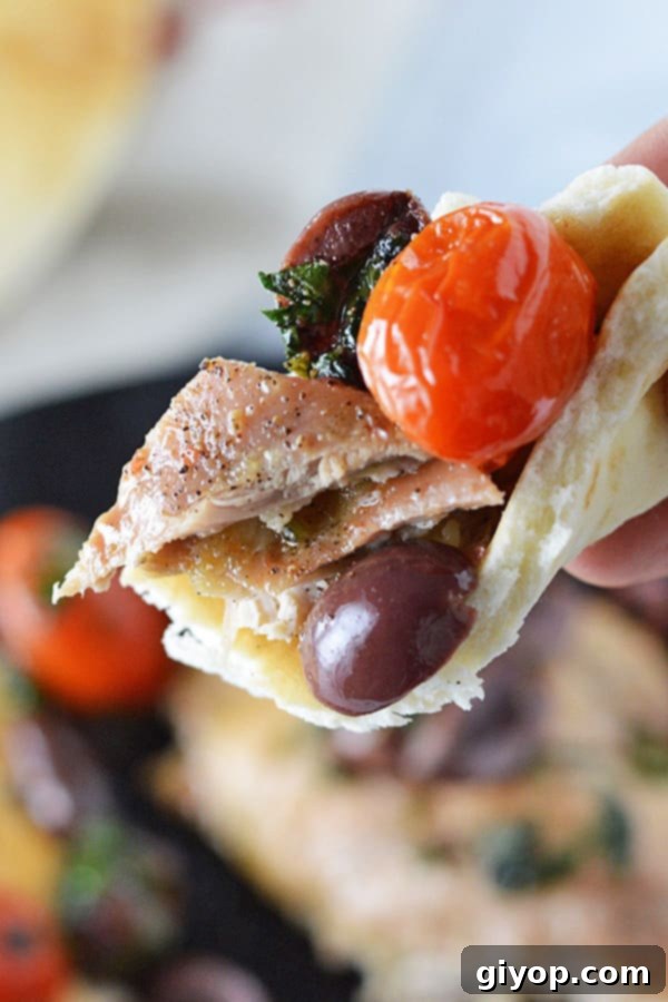 A close-up shot of a hand using a piece of warm pita bread to scoop up a mouthful of tender baked lemon pepper chicken, richly covered in the Mediterranean olive and tomato relish.