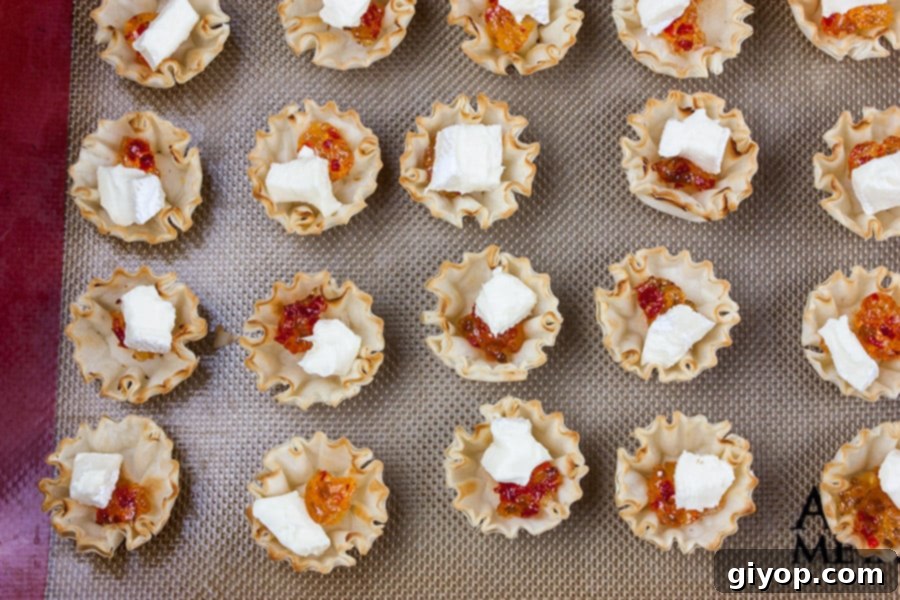 Unbaked phyllo pastry shells on a baking sheet, filled with pepper jelly and topped with small cubes of brie cheese.