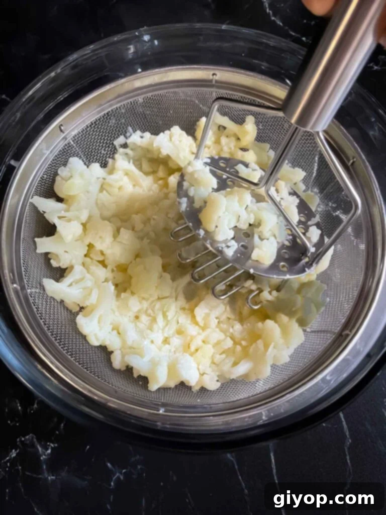 Cooked cauliflower in a fine mesh strainer being mashed with a potato masher.