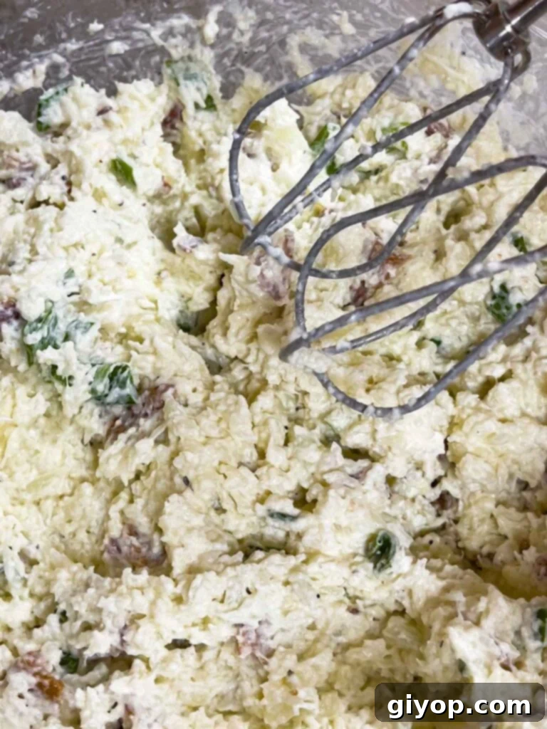 The loaded cauliflower mash in a glass bowl being mixed with an electric handheld mixer.