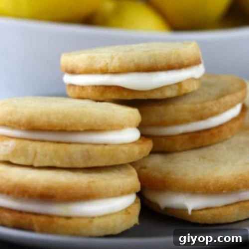 Zesty Lemon Cream Cookies 5 Lemon Sandwich Cookies stacked up on a plate with a bowl of lemons behind them.