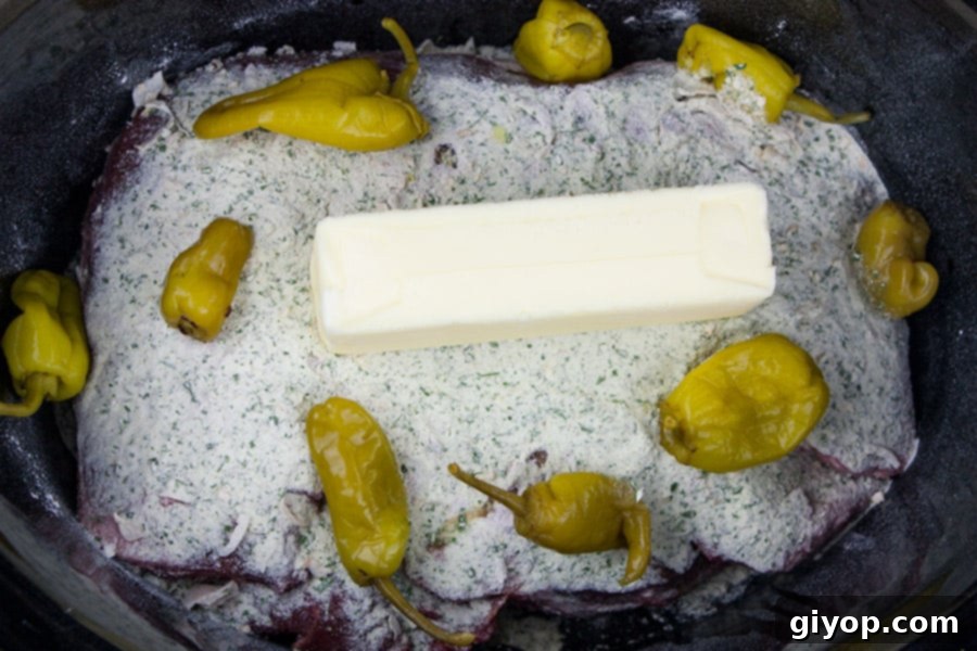Melt-in-Your-Mouth Mississippi Pot Roast 3 Chuck roast, dry ranch dressing packet, stick of unsalted butter, and pepperoncini peppers arranged in a slow cooker, ready for cooking.