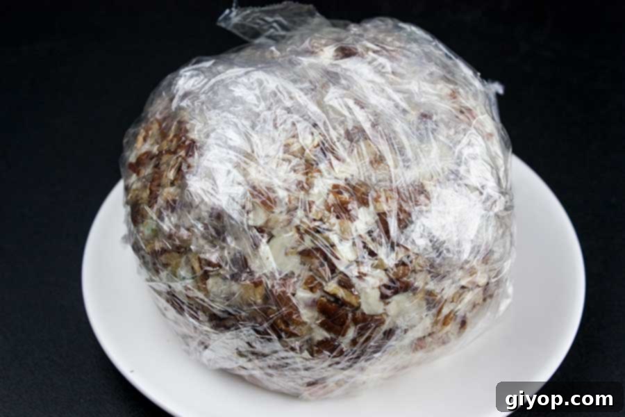 A freshly mixed cheese ball being shaped by hand on a sheet of plastic wrap covered with finely chopped pecans.