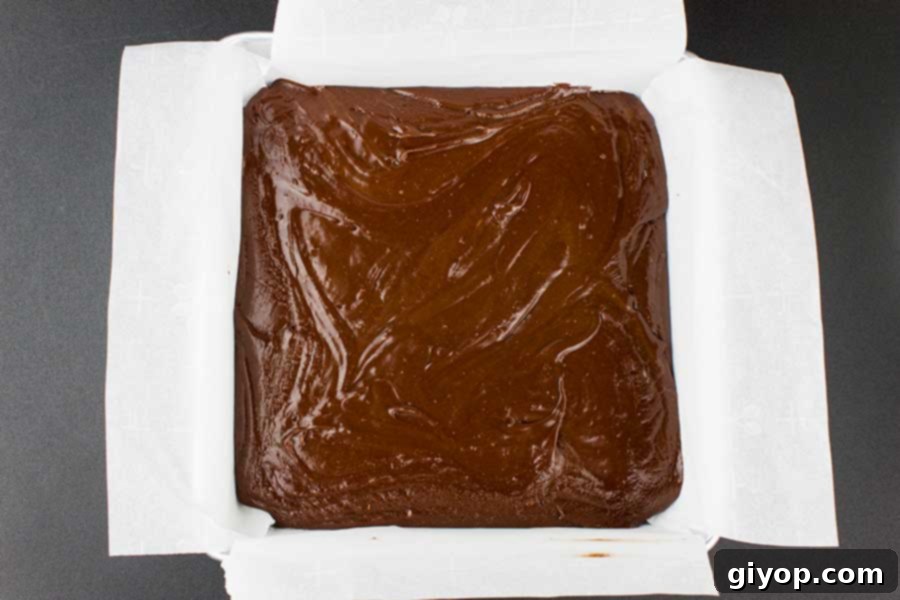 Hot fudge mixture being poured into a parchment-lined baking pan, showing its thick, glossy texture.