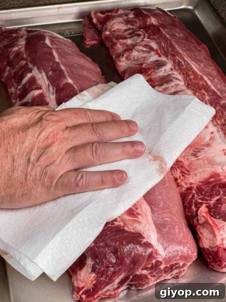 A rack of baby back ribs being patted dry with a paper towel on a cutting board, a crucial step before applying rub.