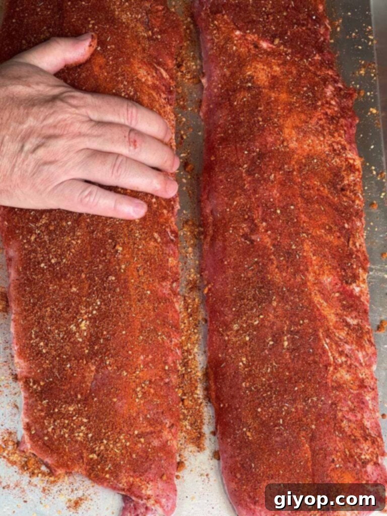 A cook applying a generous layer of Memphis style dry rub to a rack of baby back ribs, ensuring full coverage and flavor absorption.