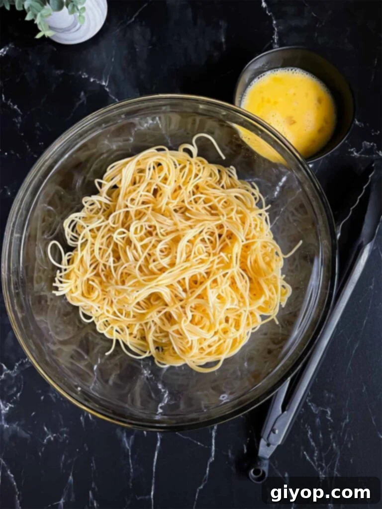 Ultimate Oven-Baked Spaghetti Delight 4 Boiled thin spaghetti in a large glass bowl with two beaten eggs in a small bowl, ready for tossing, set on a dark surface.