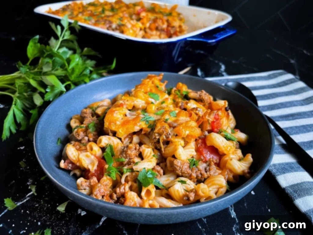 Comforting Homemade Cheesy Beefaroni 4 A serving of Cheesy Beefaroni in a dark bowl, with the casserole dish visible in the background, garnished with fresh herbs.