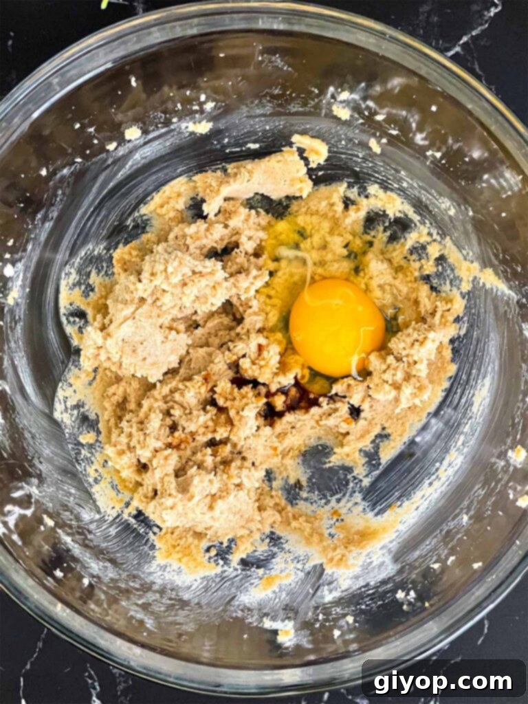 A cracked egg and a splash of vanilla extract being added to the light and fluffy creamed butter and sugar mixture for Almond Joy cookies.