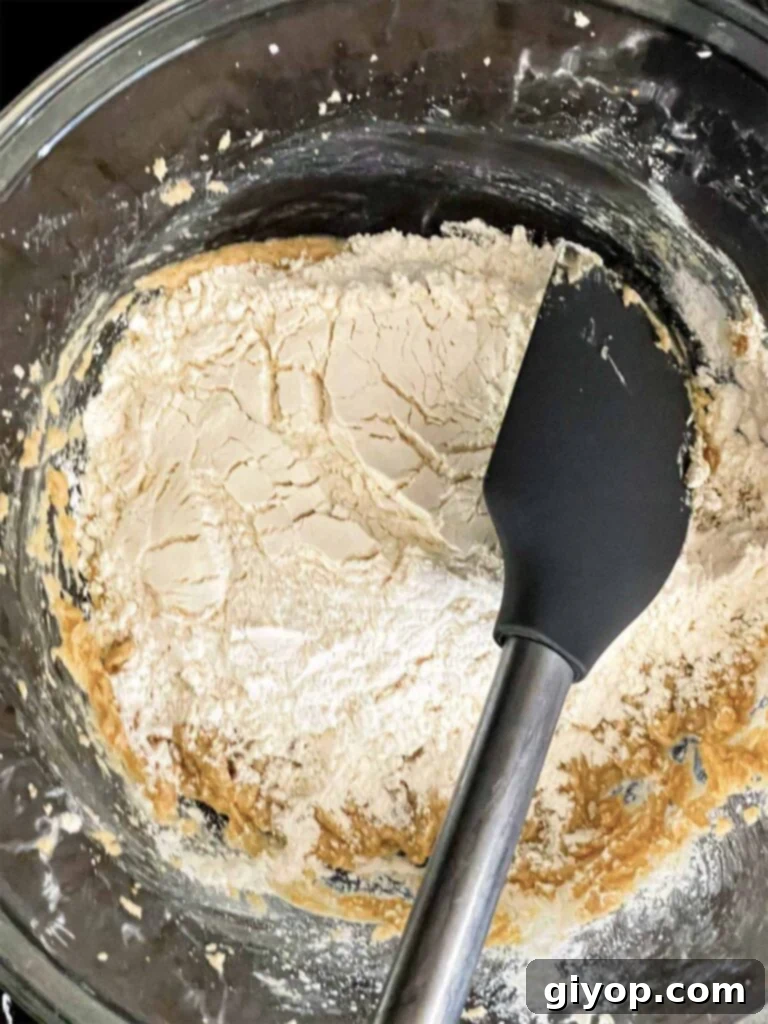 The dry flour mixture being slowly added to the wet ingredients in a large glass mixing bowl for Almond Joy cookies.