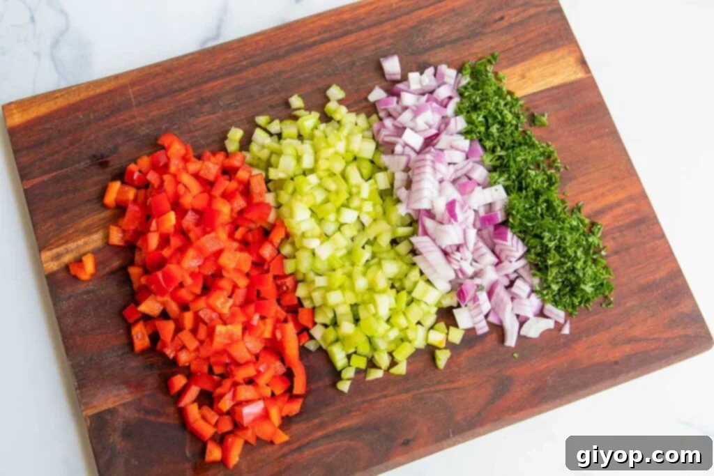 Freshly diced red bell pepper, celery, red onion, and minced parsley are displayed, ready to be added to the macaroni salad.