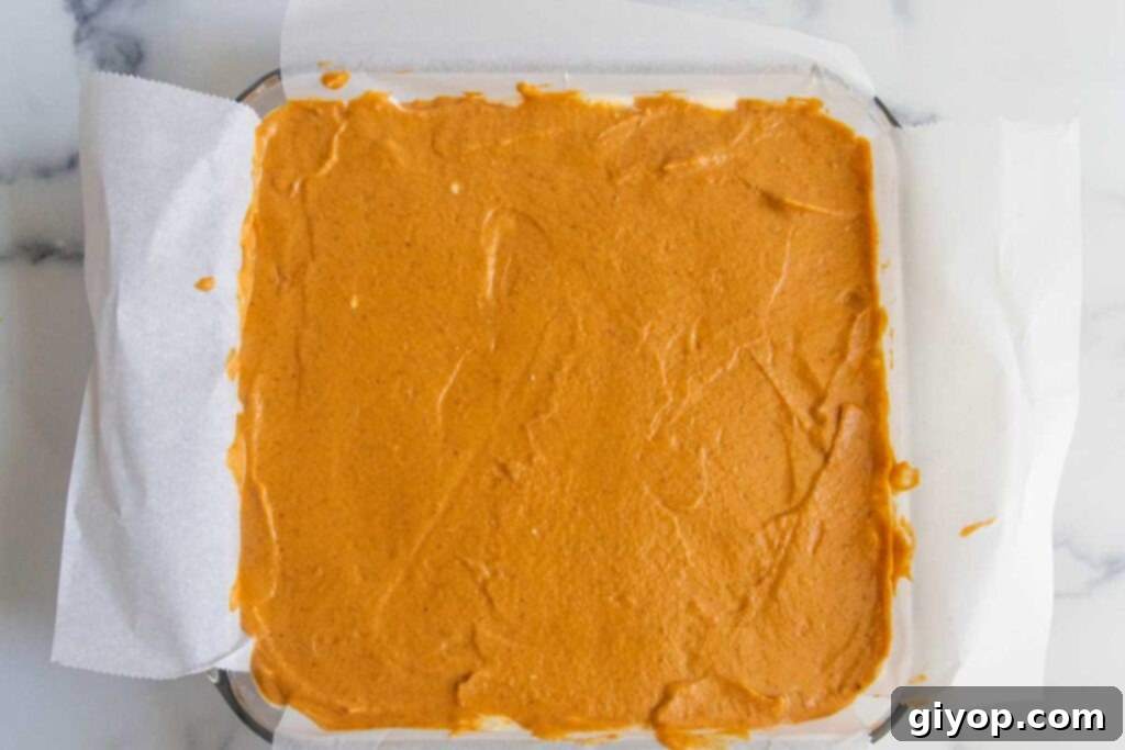 The spiced pumpkin layer is carefully spread over the plain cheesecake layer in the baking pan, creating a beautiful two-toned effect before baking.