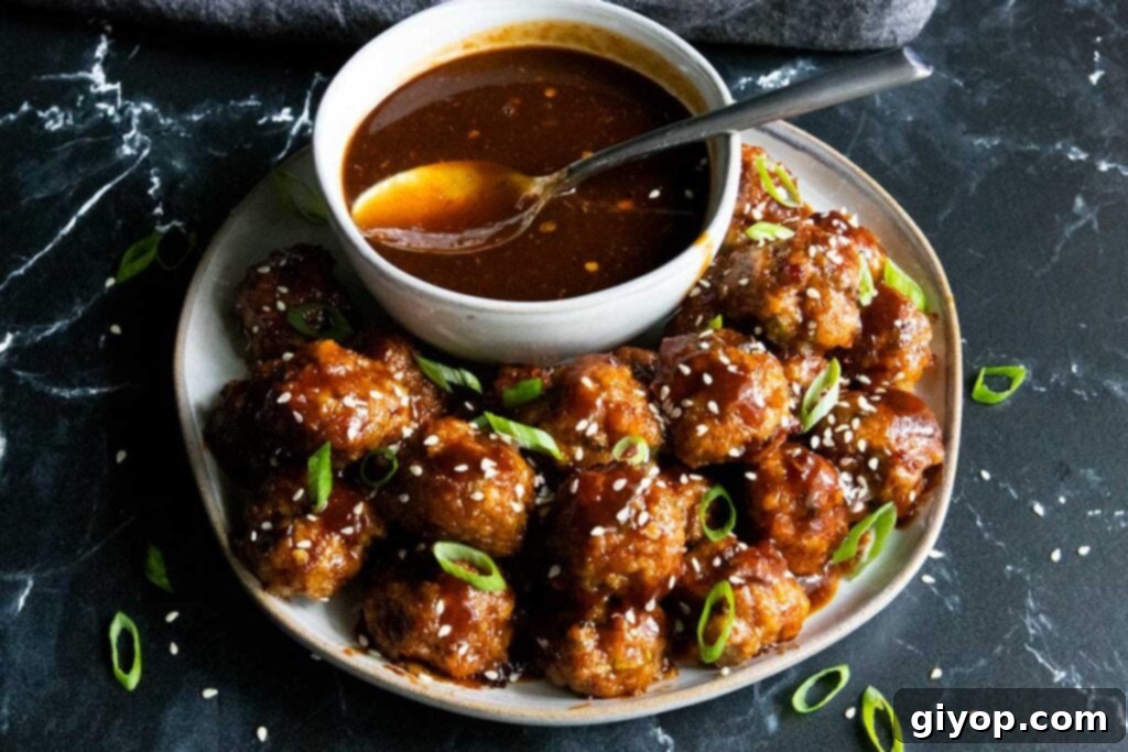 A close-up of a single Asian meatball covered in glistening sauce, garnished with sesame seeds and green onions, showcasing its delicious texture.