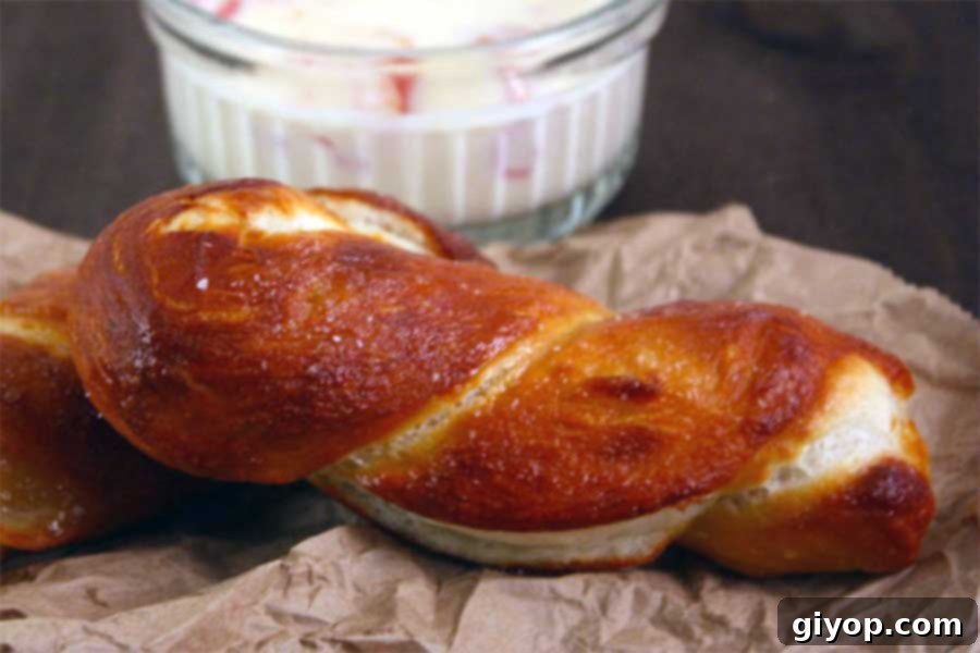 Two perfectly baked soft pretzels, one classic knot and one twisted stick, resting on brown parchment paper with a bowl of cheese dip in the background.