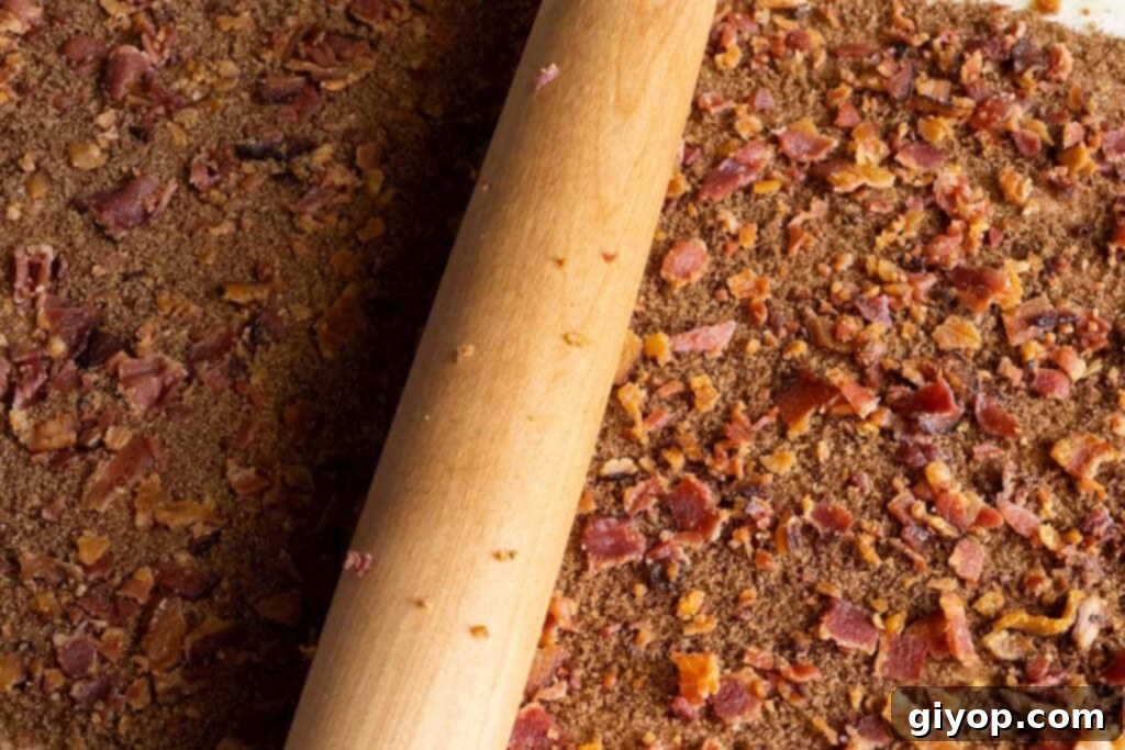 A rolling pin lightly pressing the crumbled bacon into the cinnamon-brown sugar mixture on the dough.