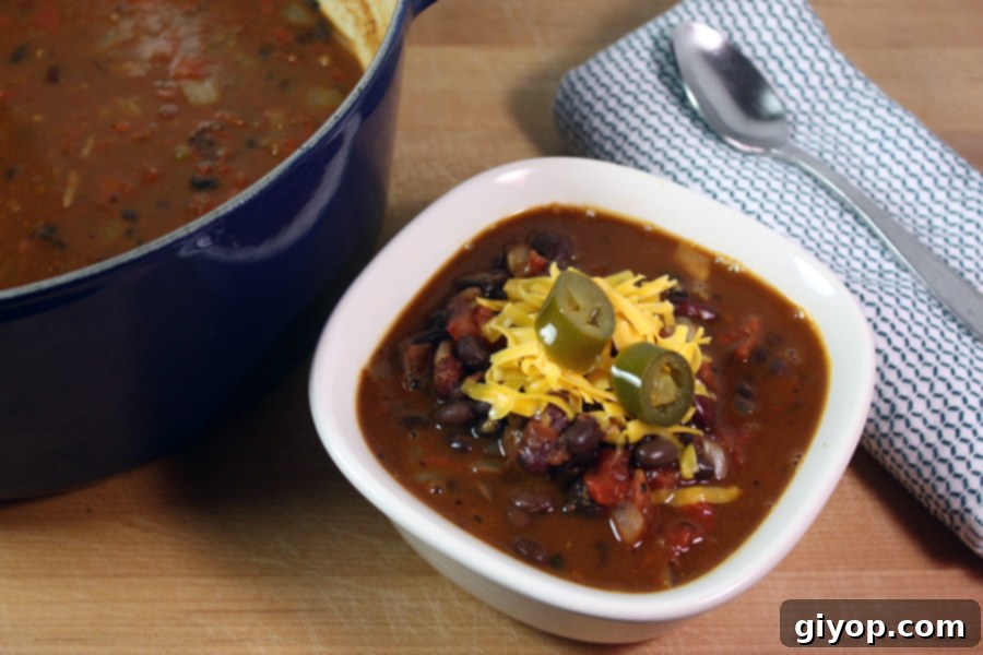 Close-up of a serving of vegetarian chili in a white bowl, garnished with generous amounts of shredded cheese and vibrant green jalapeño slices.