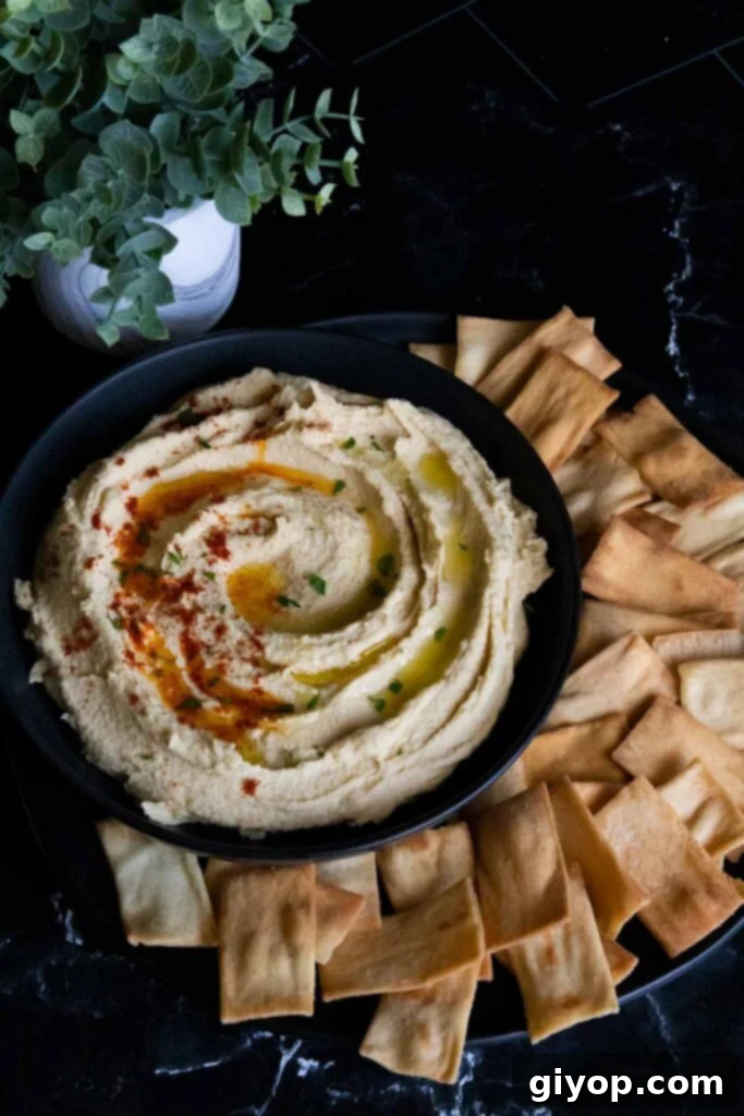 Homemade hummus in a black serving bowl, artfully garnished with a swirl of olive oil and paprika, surrounded by a variety of pita chips.