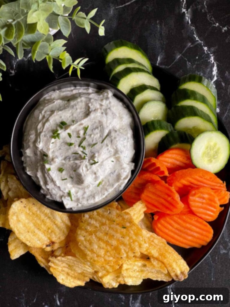 The Ultimate Chip Dip 5 A serving of homemade chip dip in a black bowl, garnished with fresh chives, surrounded by a variety of potato chips and colorful vegetables