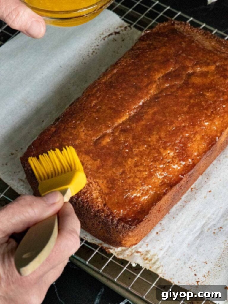 A yellow pastry brush applying melted butter over the warm apple cider doughnut cake.
