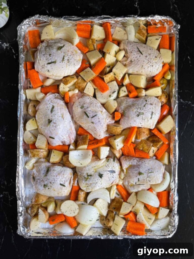 Raw chicken thighs arranged skin-side up amidst seasoned potatoes, carrots, onions, and garlic on a baking sheet, ready for the oven.