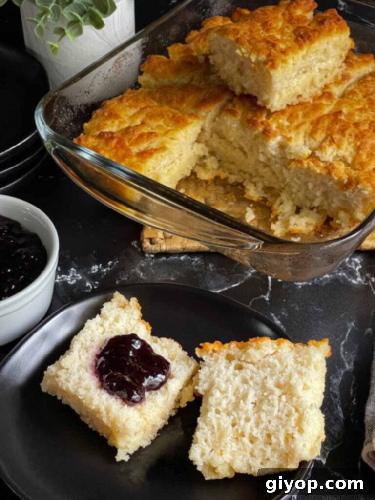 A butter swim biscuit, perfectly golden and cut in half, showcasing its fluffy interior, with a dollop of blackberry jam on one side, resting on a sleek black plate.