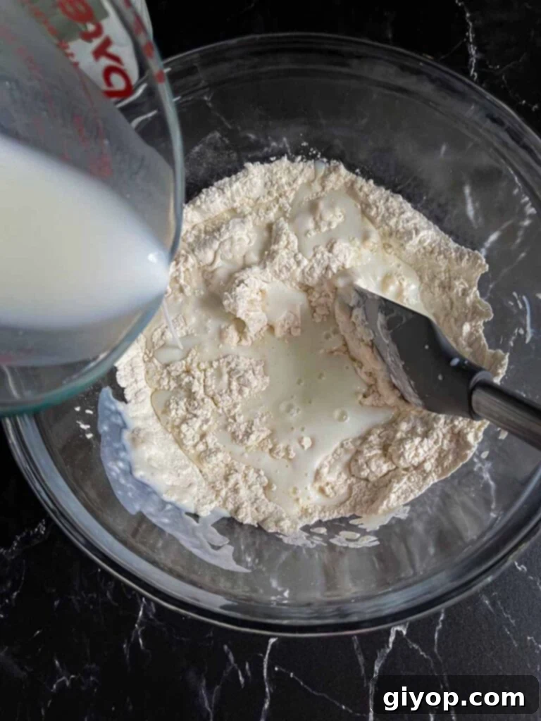 Buttermilk being stirred into the dry ingredients in a clear glass mixing bowl, forming a shaggy dough.