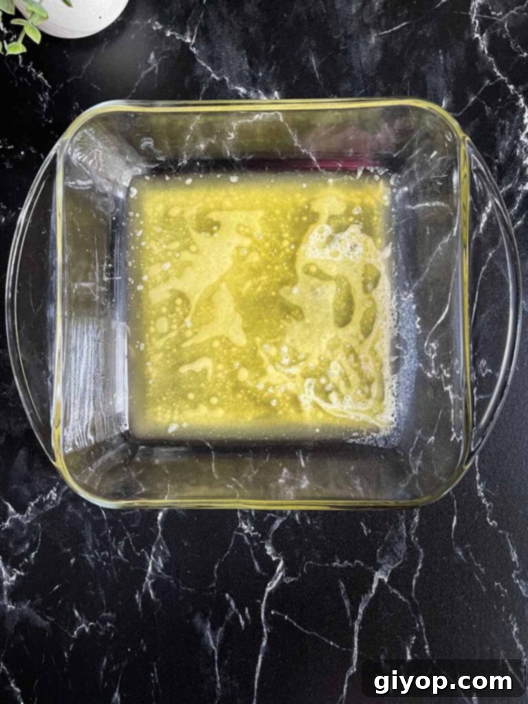 A square glass baking dish filled with a shimmering pool of melted butter, ready for the biscuit dough.