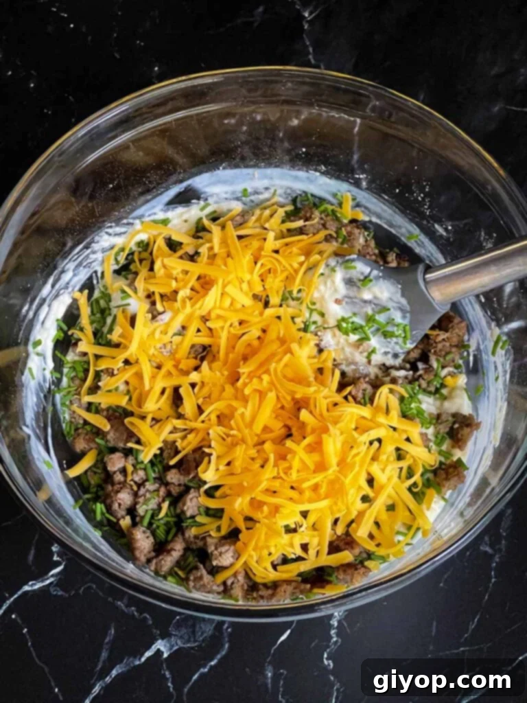 Cooked sausage, shredded cheddar cheese, and fresh chives being gently folded into the muffin batter with a silver spatula.