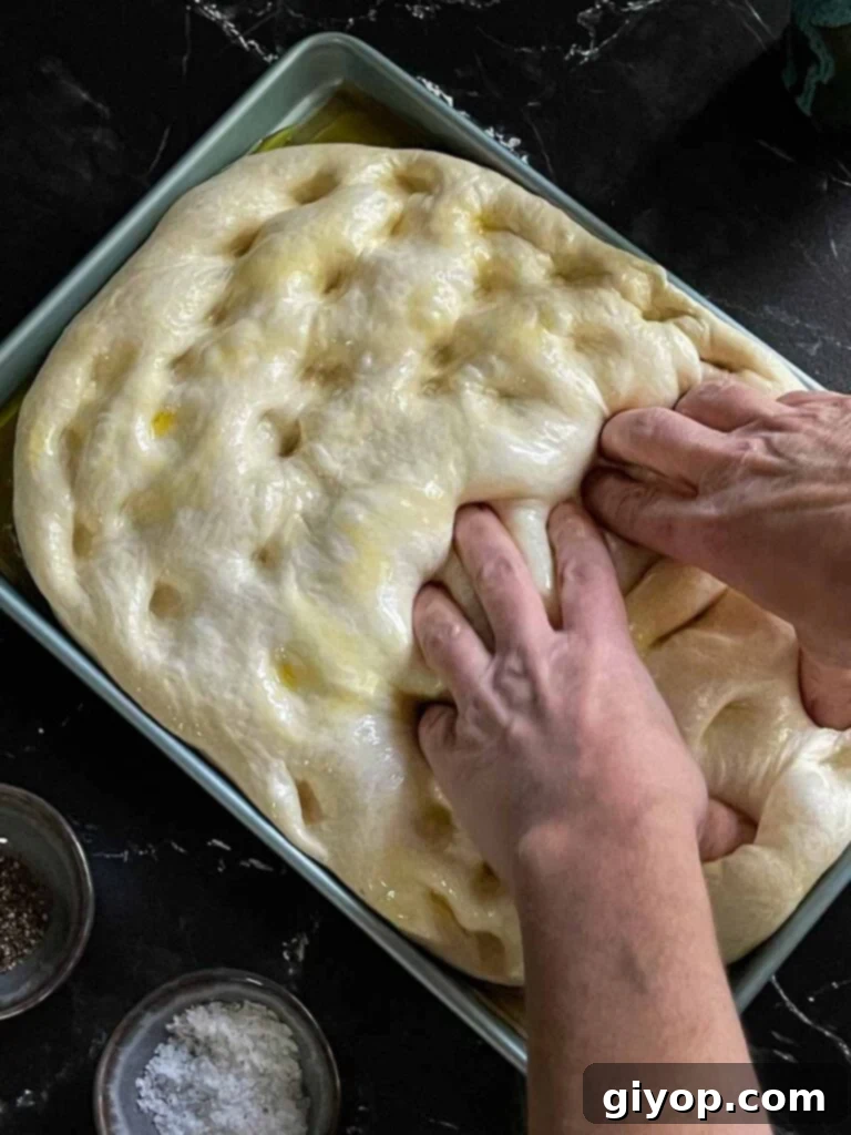 Fingers pressing into the risen focaccia dough to create characteristic dimples, shaping it to the pan.