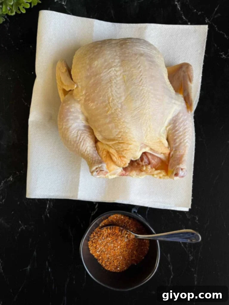 Raw whole chicken on paper towels being patted dry, with a small ramekin of spice rub on a dark surface.