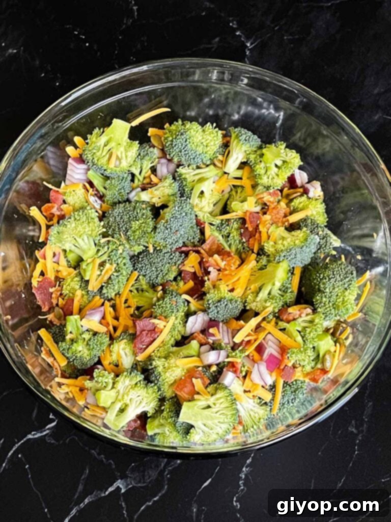 Broccoli salad ingredients including fresh broccoli, red onion, cheese, bacon, and pumpkin seeds, are tossed together in a large glass mixing bowl.