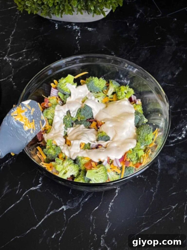 The rich, creamy dressing is being poured over the vibrant broccoli salad ingredients in a clear glass mixing bowl, ensuring every piece is coated.