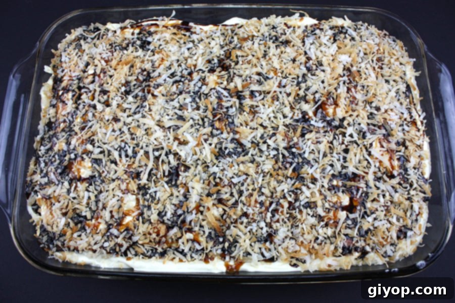 A partially assembled Samoa Cake in a glass baking dish, showing the first cookie layer topped with creamy coconut pudding, and drizzled with caramel and chocolate sauces.