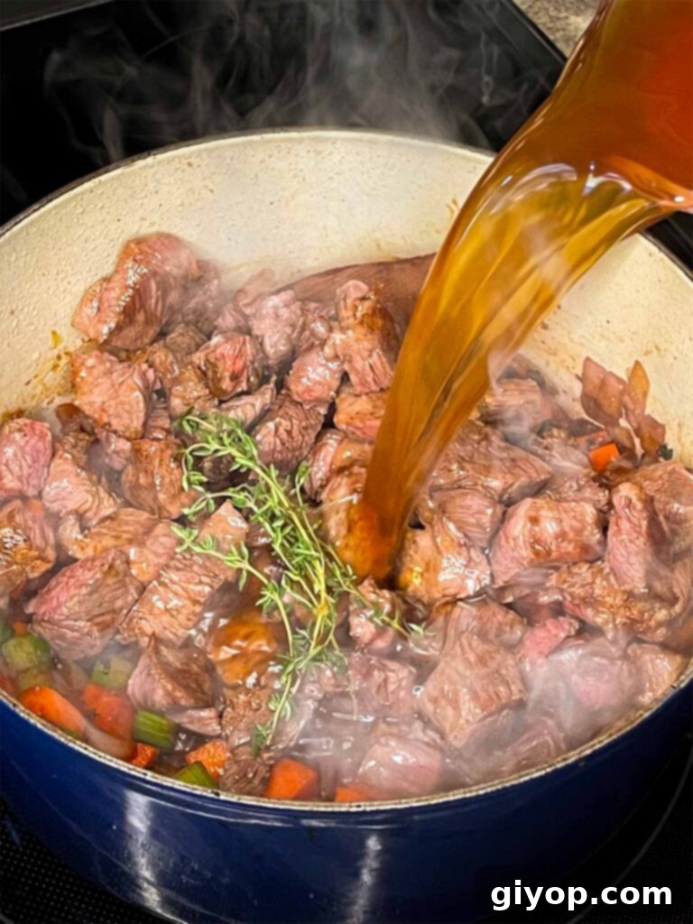 Pouring rich beef broth into the Dutch oven with browned beef and sautéed vegetables, setting the stage for simmering.
