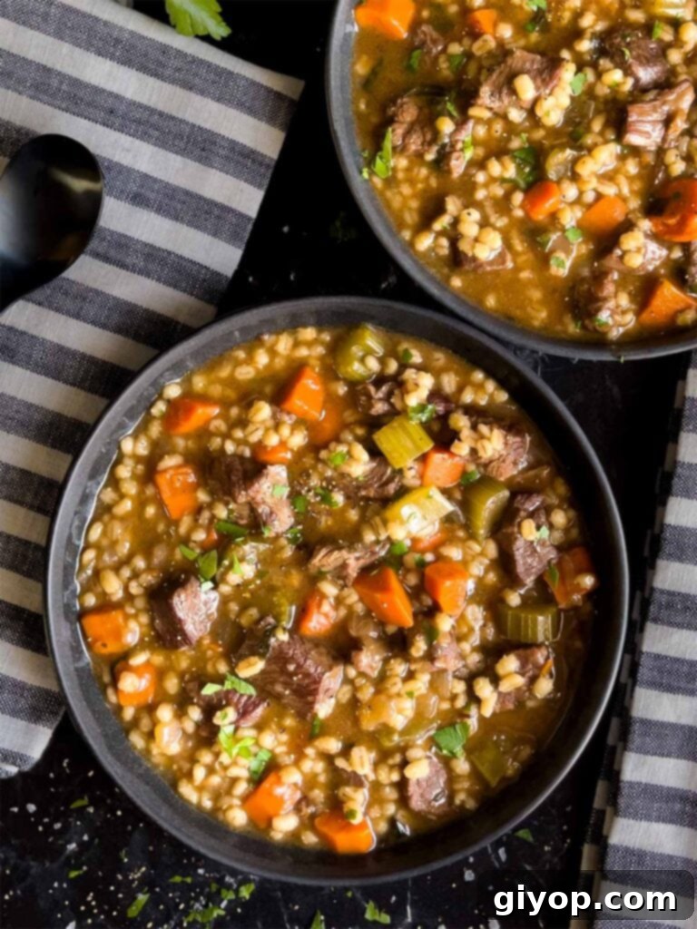 Two dark bowls of beef barley soup, garnished with fresh parsley, placed next to striped napkins on a rustic wooden surface.