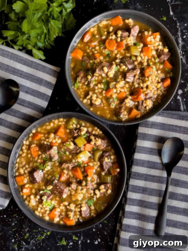 Two cozy bowls of beef barley soup, garnished with fresh parsley, paired with striped cloth napkins on a textured wooden table.