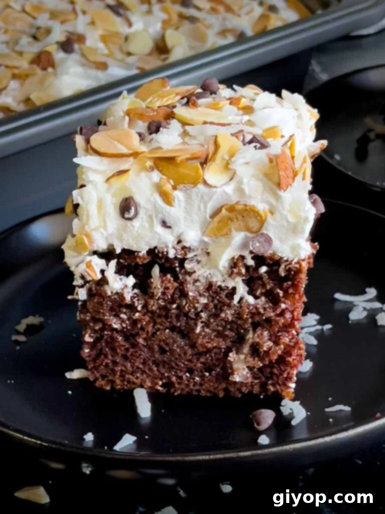 Slice of Almond Joy cake on a dark plate, showcasing layers of chocolate cake, coconut sauce, whipped topping, almonds, and chocolate chips.
