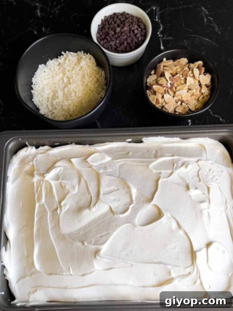 Whipped topping being spread smoothly over the cooled Almond Joy cake, with bowls of coconut, almonds, and chocolate chips in the background.