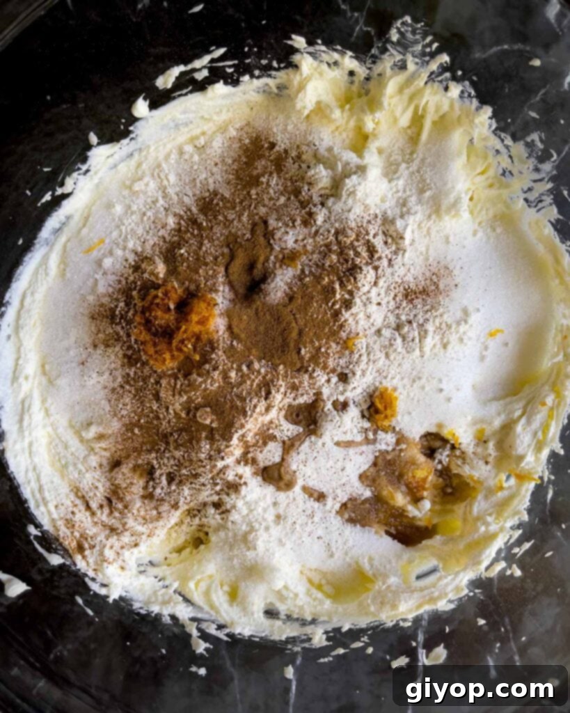 A mix of granulated sugar, ground cinnamon, all-purpose flour, fresh orange zest, vanilla extract, and kosher salt being added to the creamy cannoli cheesecake mixture in a glass bowl.