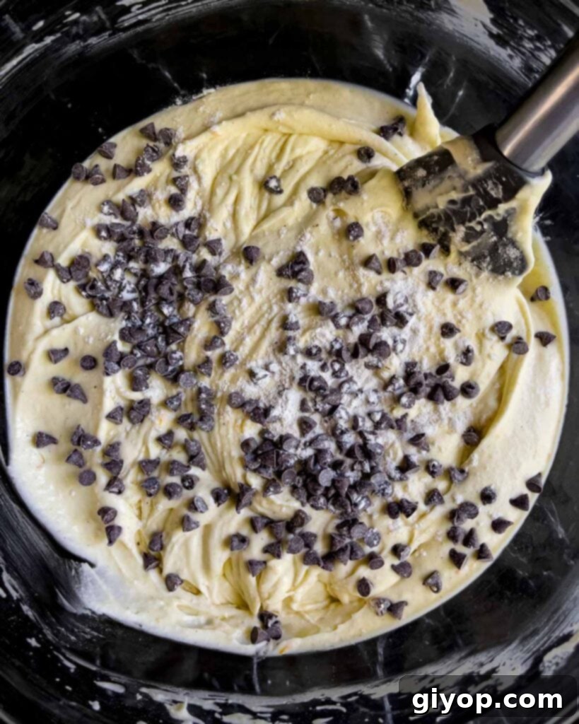 Mini chocolate chips being carefully folded into the smooth cannoli cheesecake batter in a glass bowl, ensuring even distribution.