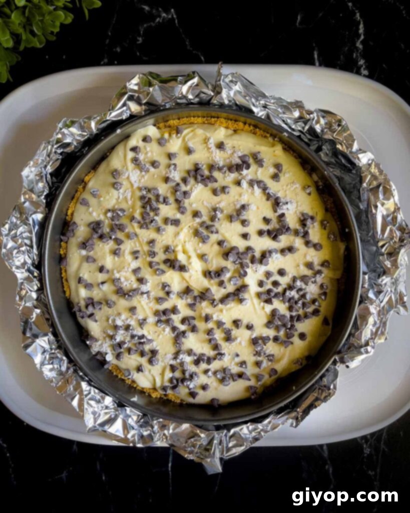 The cannoli cheesecake in its springform pan, securely wrapped with aluminum foil, carefully placed inside a larger roasting pan for a water bath.