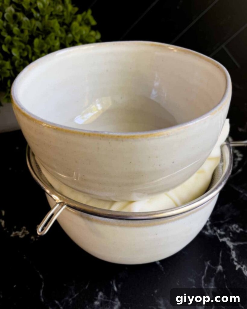 Ricotta cheese being prepared for draining, placed in cheesecloth within a strainer, which is set over a bowl. Another bowl is positioned on top to weigh it down, encouraging liquid extraction.