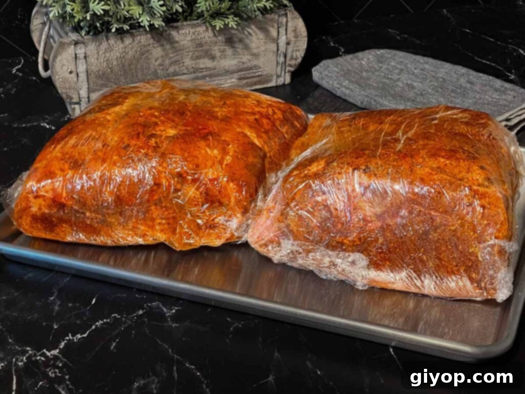 Two pork butts generously coated with rub and wrapped in plastic wrap, ready for smoking.