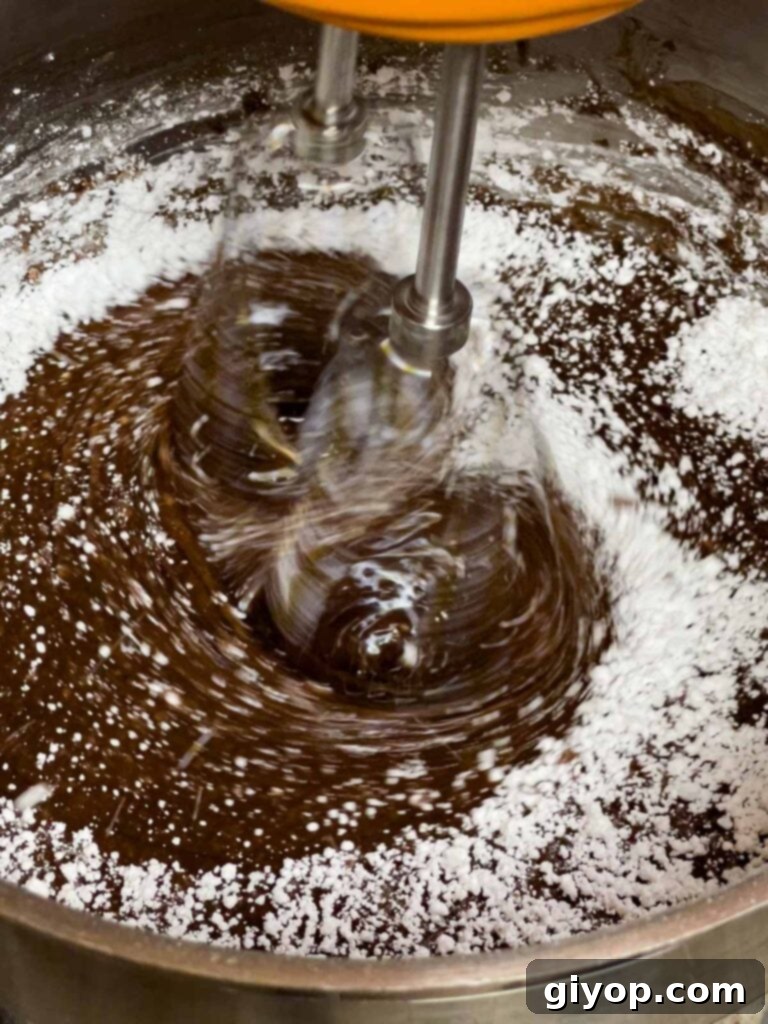 Powdered sugar being beaten into the warm chocolate frosting for the Mississippi Mud Cake with an electric mixer, creating a smooth consistency.