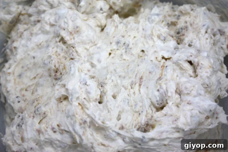 A clear mixing bowl containing softened cream cheese, powdered sugar, vanilla extract, and salt, being skillfully whipped to a smooth, airy consistency with an electric mixer.